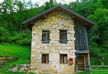 نمای بیرونی یک خانه روستایی دو طبقه در جنگل که دیوارهای آن از سنگ‌های لاشه طبیعی و کوهی به رنگ کرم و قهوه‌ای ساخته شده است.