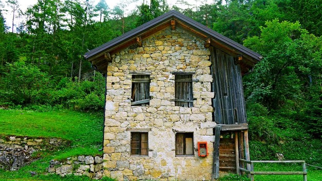 نمای بیرونی یک خانه روستایی دو طبقه در جنگل که دیوارهای آن از سنگ‌های لاشه طبیعی و کوهی به رنگ کرم و قهوه‌ای ساخته شده است.