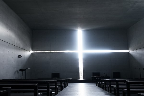 نمای داخلی کلیسای نور با دیوارهای بتنی خاکستری و شکافی به شکل صلیب در دیوار روبرو که نور خورشید را به صورت درخشان به داخل فضای تاریک هدایت میکند.