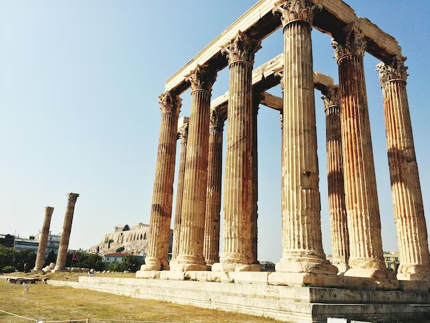 ستونهای عظیم و سنگیِ به جامانده از یک معبد باستانی در یونان (آتن) با سرستونهای کورینتی زیر آسمان صاف؛ نمادی از پایداری و هنر در معماری کهن.