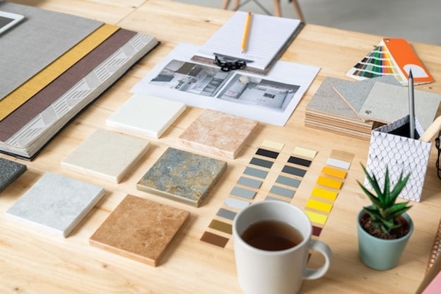 مجموعه‌ای از تایل‌های کوچک سنگ‌های ساختمانی مختلف مانند مرمریت و تراورتن در کنار پالت رنگ و نقشه‌های طراحی روی میز چوبی.