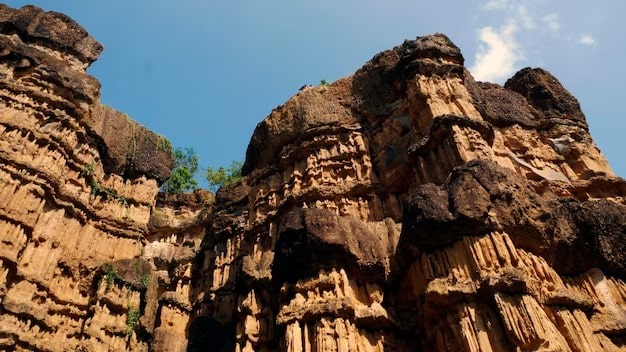 نمای زیرین از صخره‌های عظیم و لایه‌لایه رسوبی با رنگ قهوه‌ای و کرم که دچار فرسایش طبیعی شده‌اند زیر آسمان صاف.