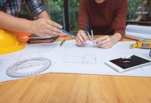 مهندس معمار در حال بررسی و اندازه‌گیری نقشه‌های فنی ساختمان با پرگار و ابزار مهندسی روی میز کار.