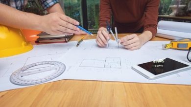 مهندس معمار در حال بررسی و اندازه‌گیری نقشه‌های فنی ساختمان با پرگار و ابزار مهندسی روی میز کار.