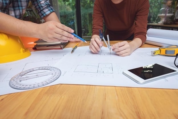 مهندس معمار در حال بررسی و اندازه‌گیری نقشه‌های فنی ساختمان با پرگار و ابزار مهندسی روی میز کار.