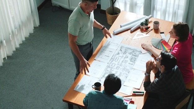 تیم مهندسی و طراحی در حال بحث و تبادل نظر روی انواع نقشه‌های معماری و پلان‌های ساختمانی دور میز کار.